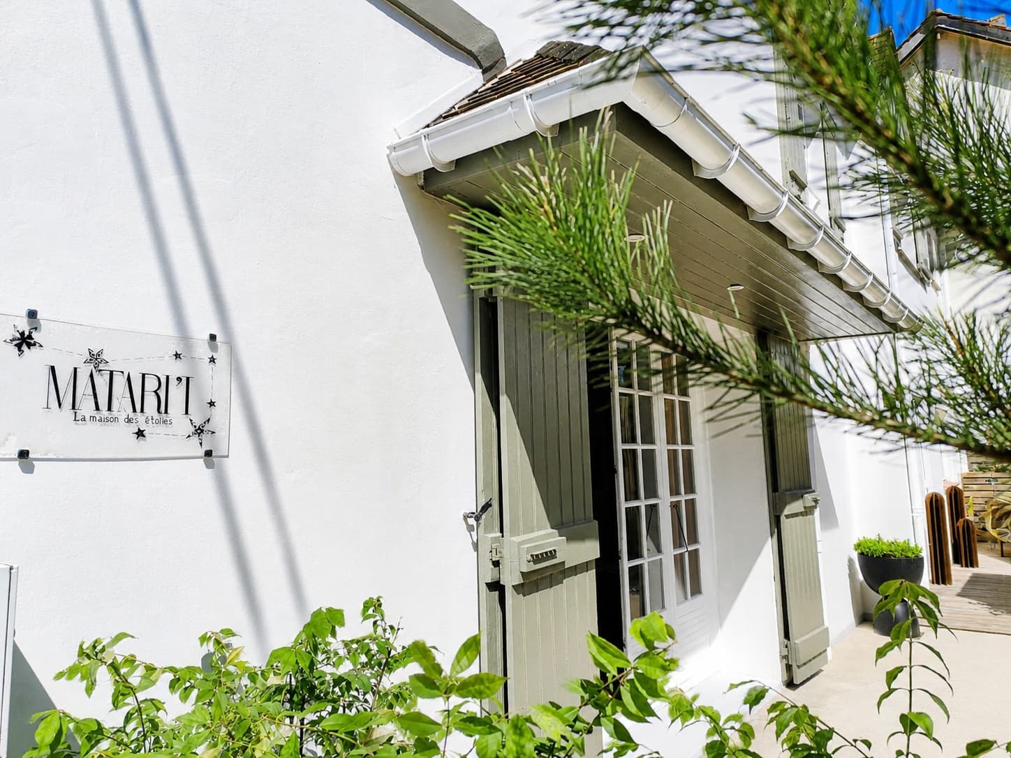 Façade blanche d’une maison accompagnée, volets gris-vert et branche de pin maritime au premier plan