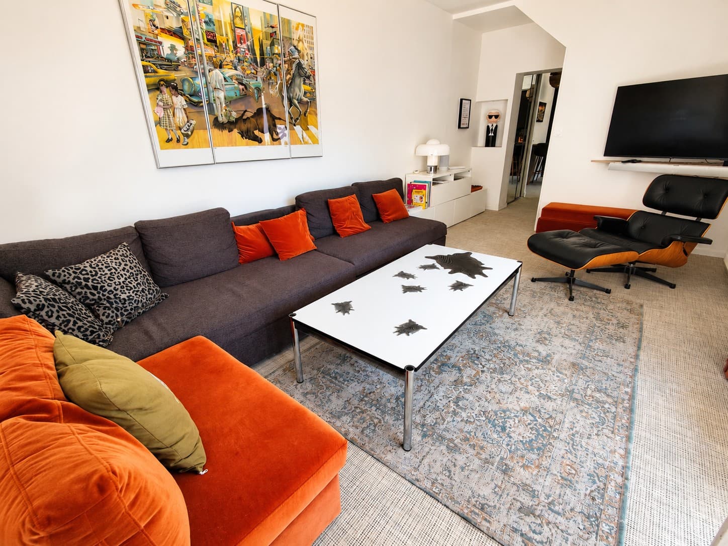 Séjour lumineux : canapé d’angle gris et velours orangé, table basse blanche à pieds chromés, fauteuil en cuir noir sur un tapis aux motifs anciens